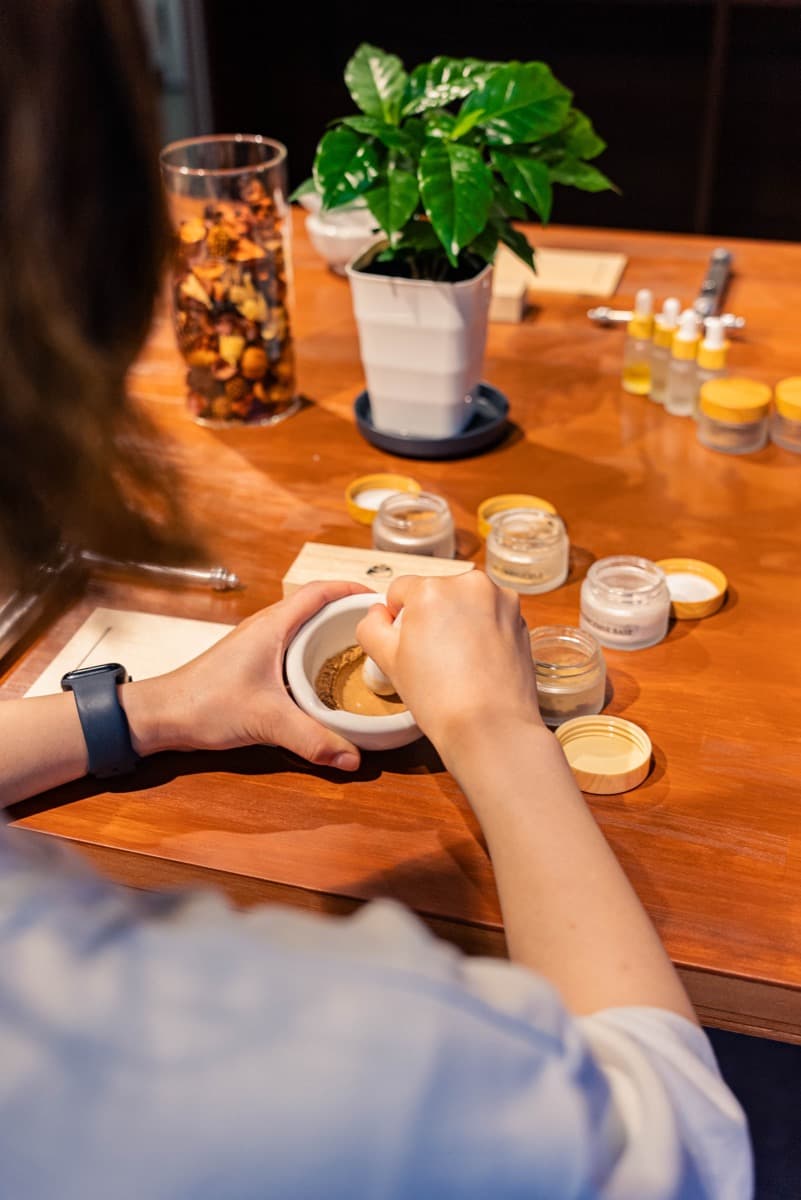 Hands grinding fragrance in a mortar, with jars of base scents like sandalwood and frankincense